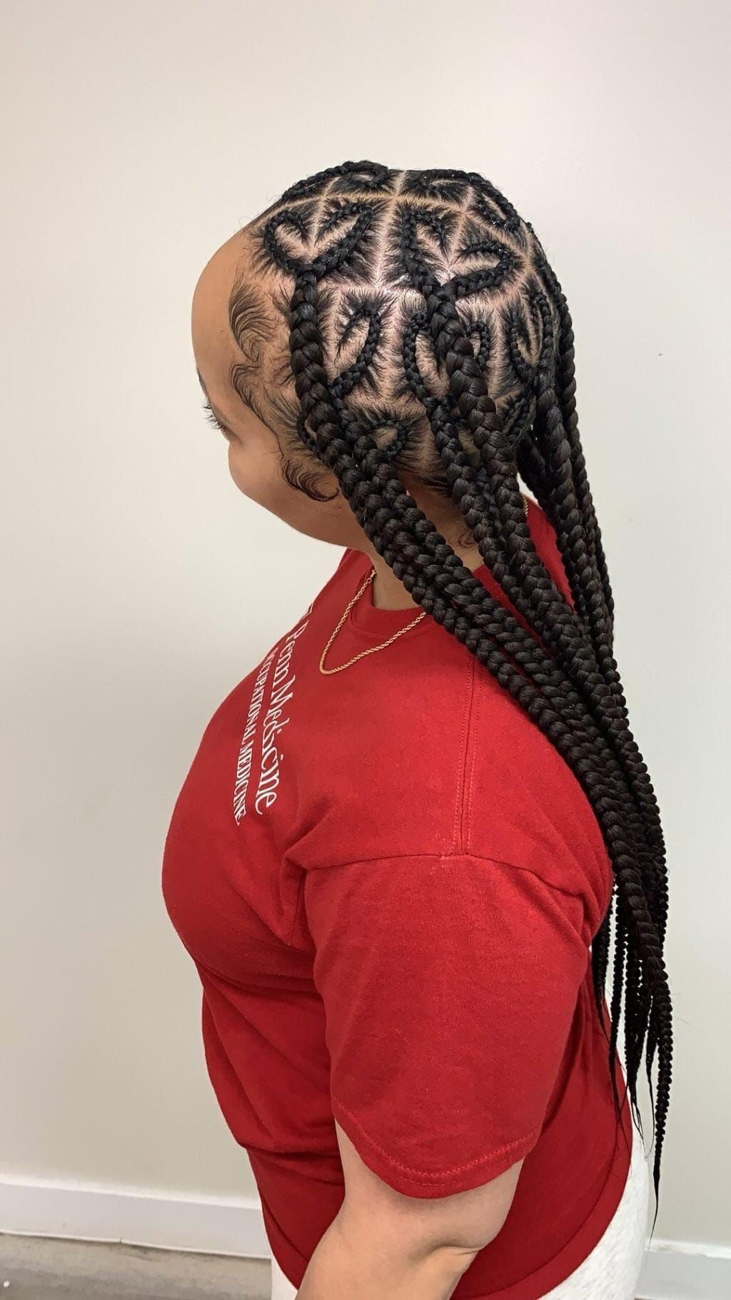 A person in a red shirt shows off a hairstyle featuring intricate, heart-shaped cornrow designs on their scalp.