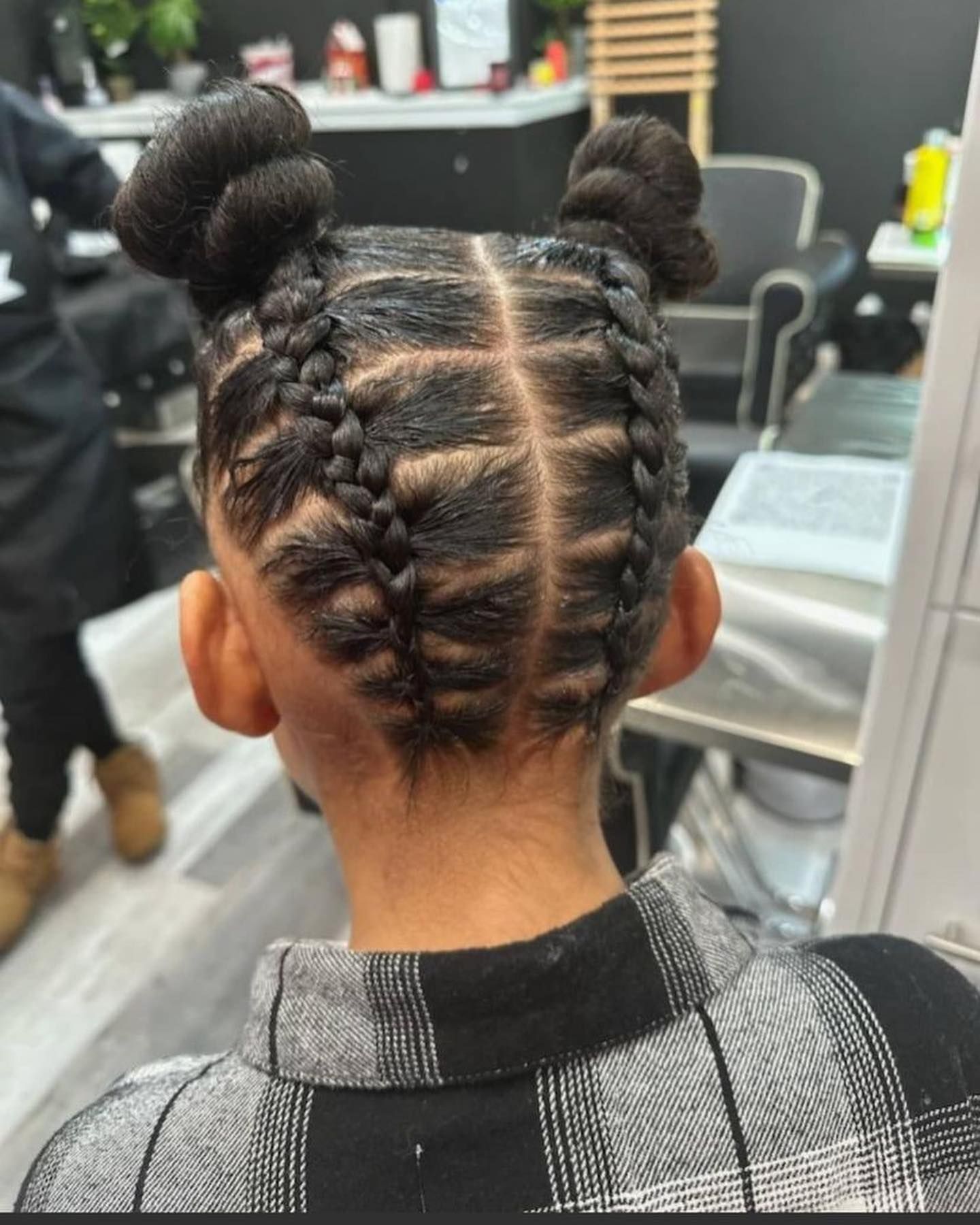 A person with dark hair styled in two symmetric cornrow braids leading into two high, circular buns in a salon setting.