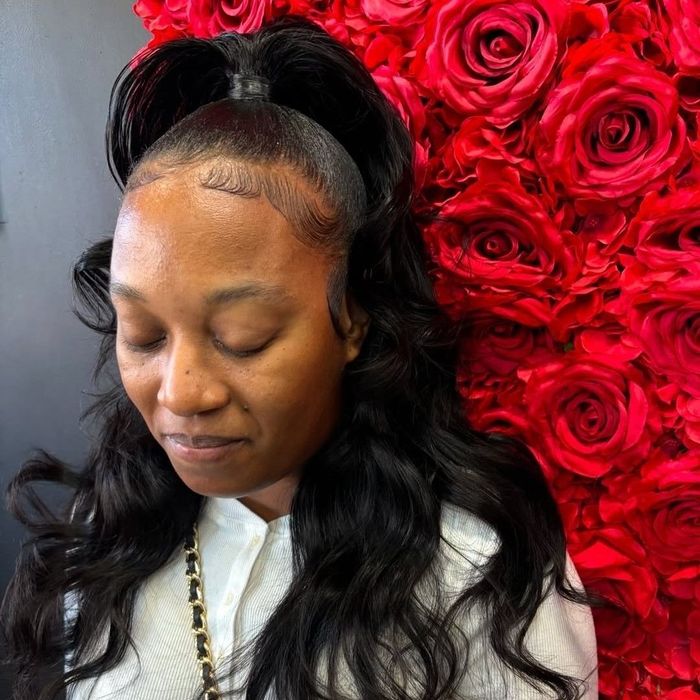 A person with styled long dark hair and a half-up ponytail standing in front of a red rose flower wall.