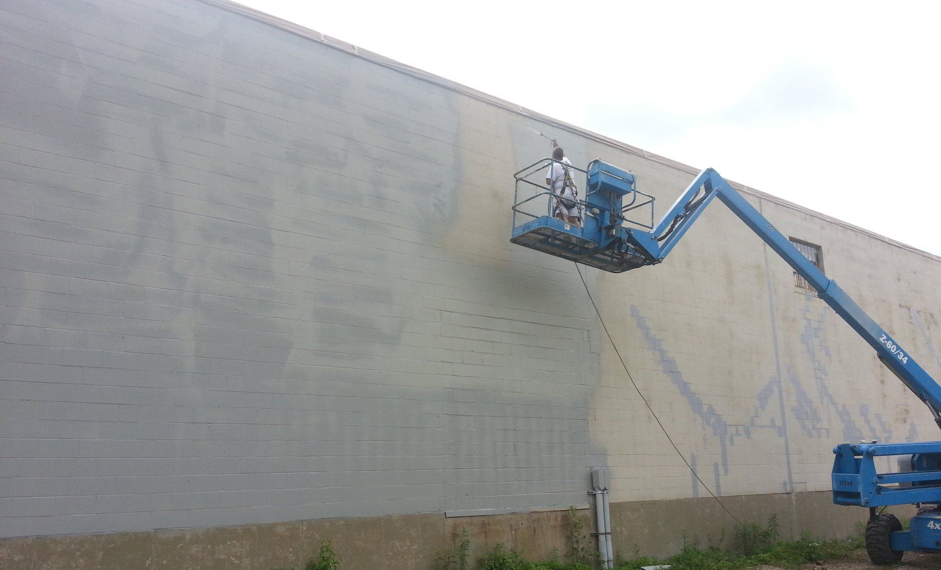 Worker in blue lift painting a large gray wall outdoors