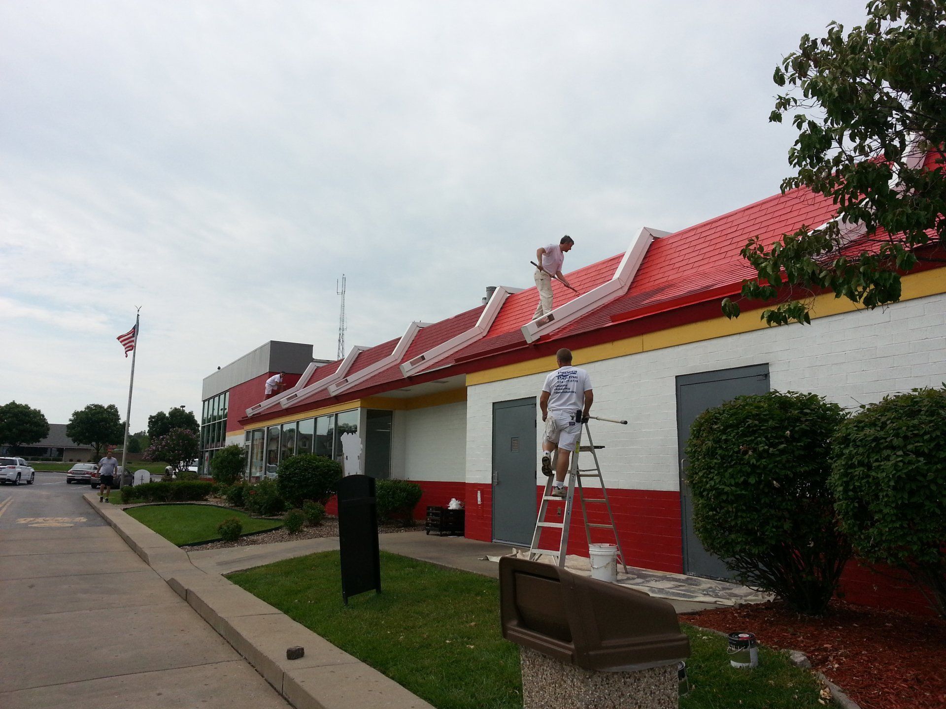 Street view of a building with a red roof, hedges, and a person on a ladder painting the exterior