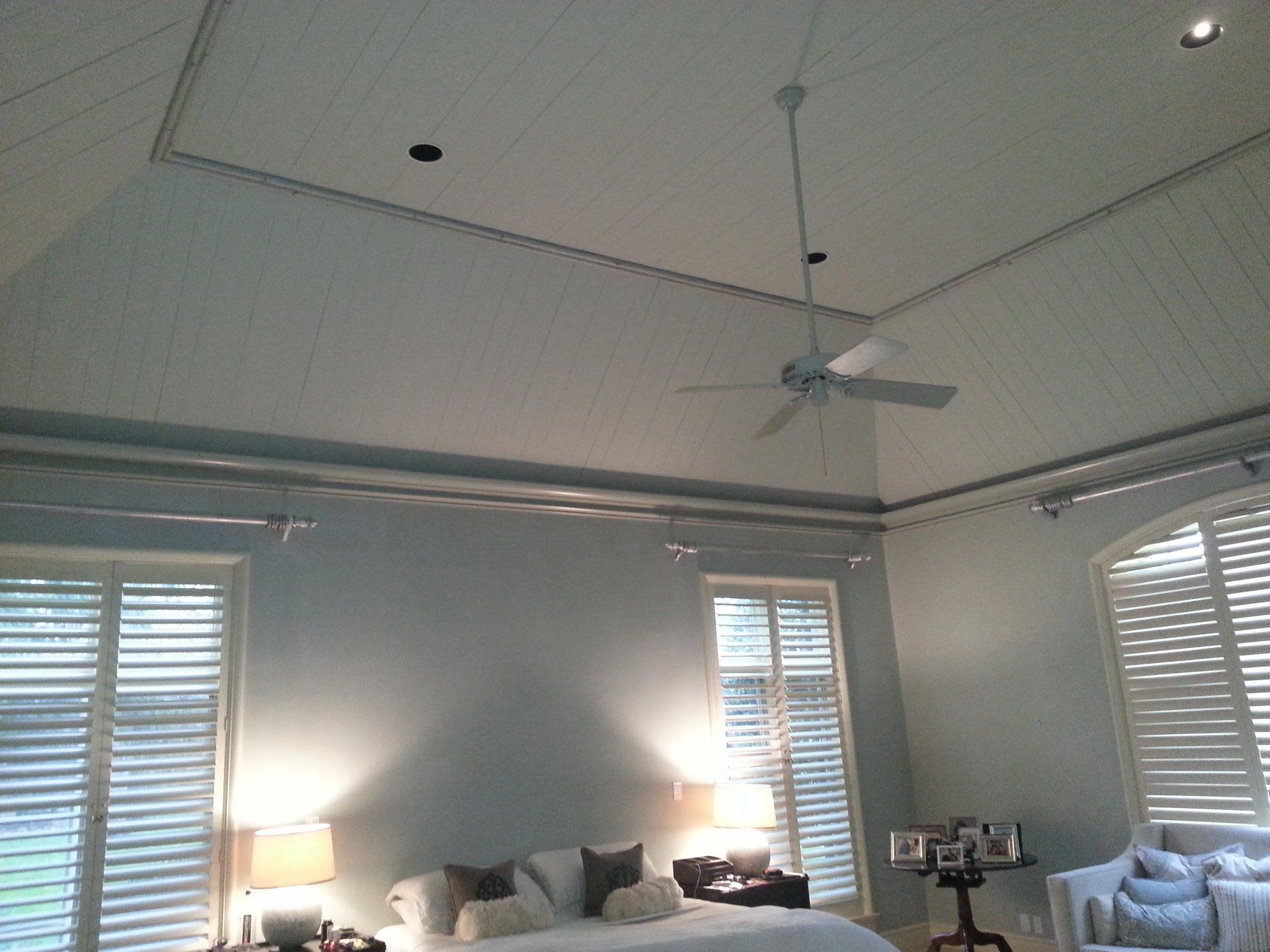 Bright bedroom with white walls, ceiling fan, lit bedside lamp, and shuttered windows.
