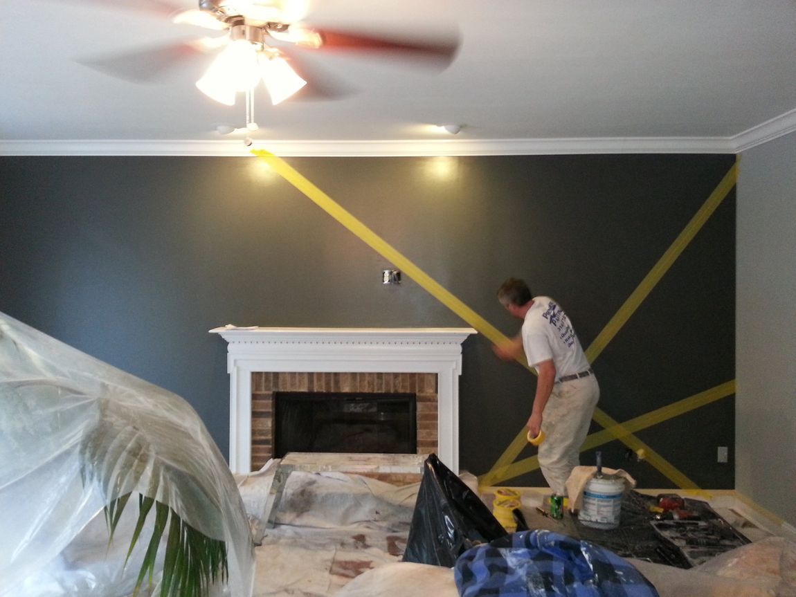 Worker taping off a dark gray living room wall around a white fireplace during painting prep