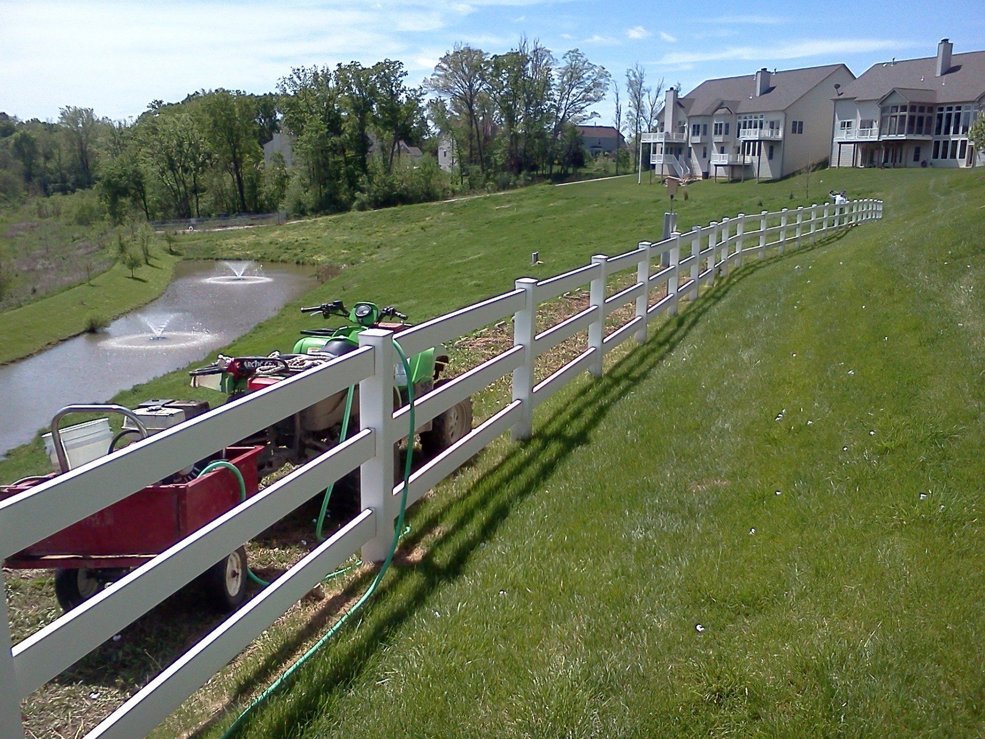White fence along a grassy hillside beside a pond and houses on a sunny day