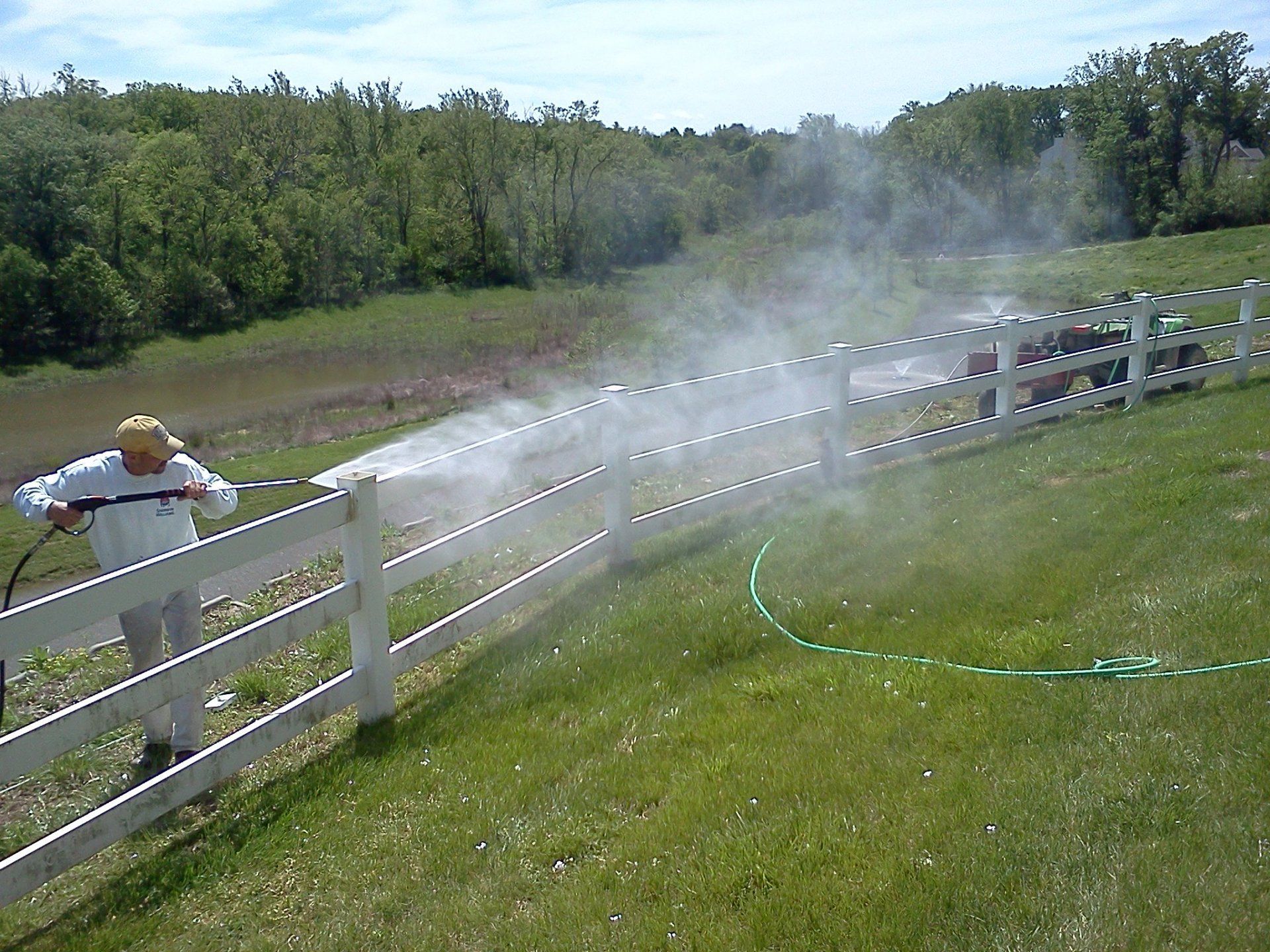 Person spraying water on a white fence in a grassy field with mist drifting over the hill