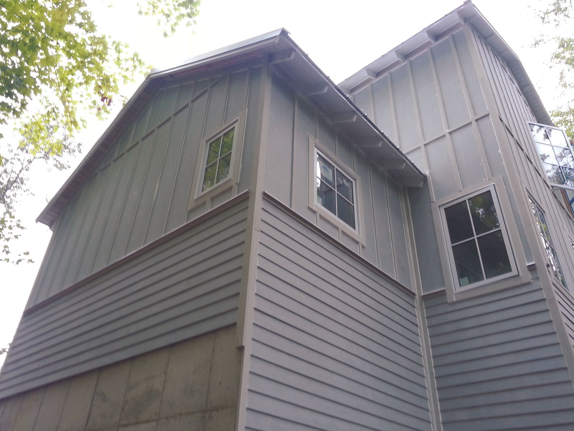Tall gray-sided house with multiple windows viewed from below among trees