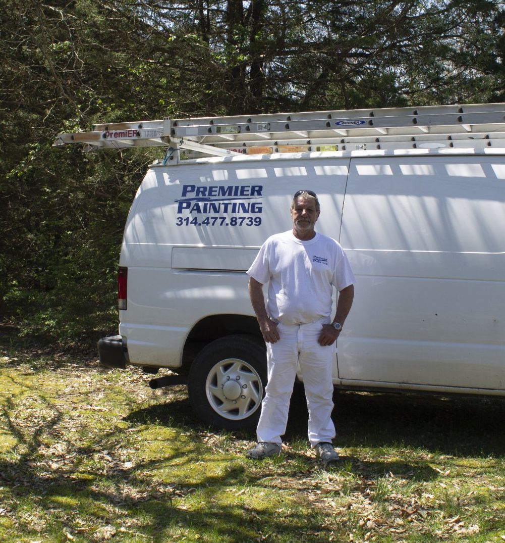 Man in white work clothes stands beside a white service van with “Premier Painting” logo in a wooded area.