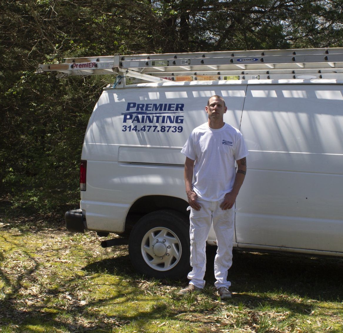 Man standing beside a white Premier Painting van in a grassy yard, holding a ladder overhead.