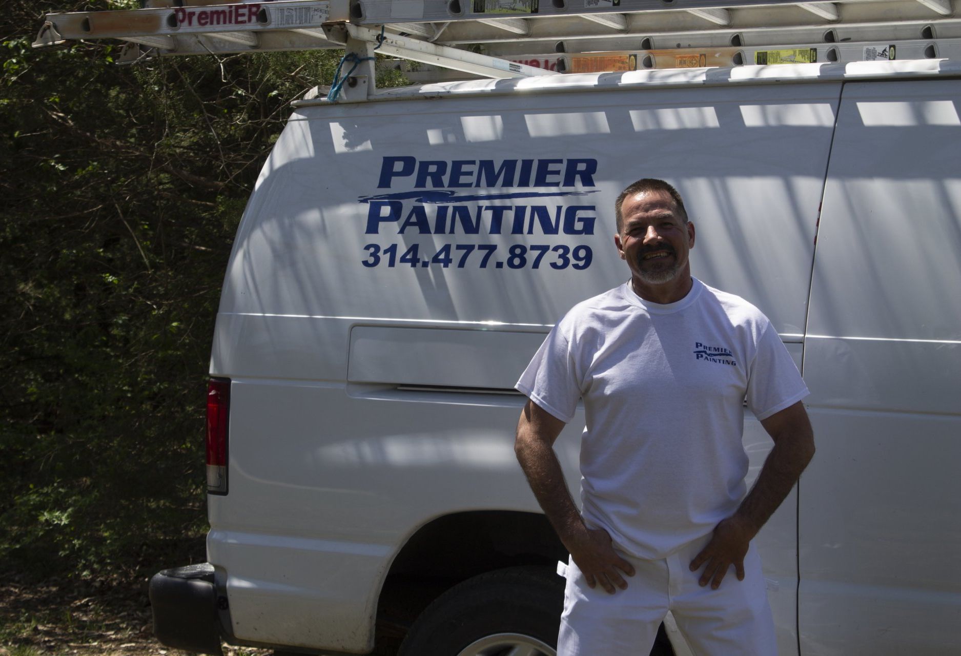 Man in white work clothes standing by a white van labeled “Premier Painting” with phone number 314-477-8739