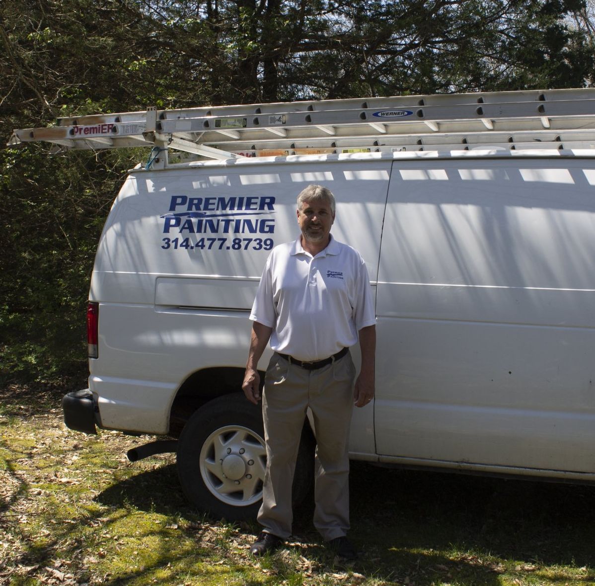 Man standing beside a white Premier Painting van with ladder on top in a wooded area