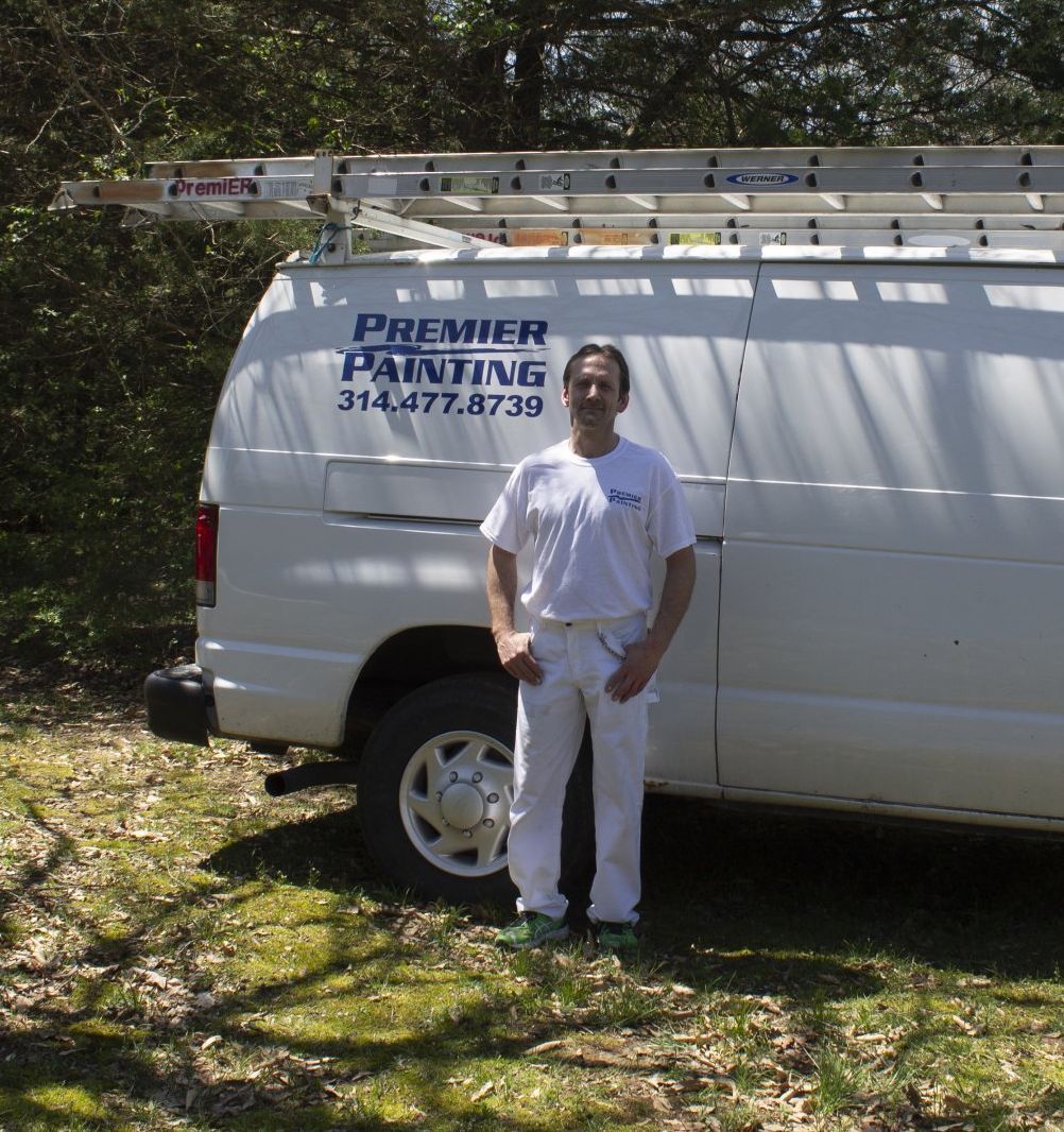 Man in white work clothes standing beside a white service van with a ladder on top in a wooded yard