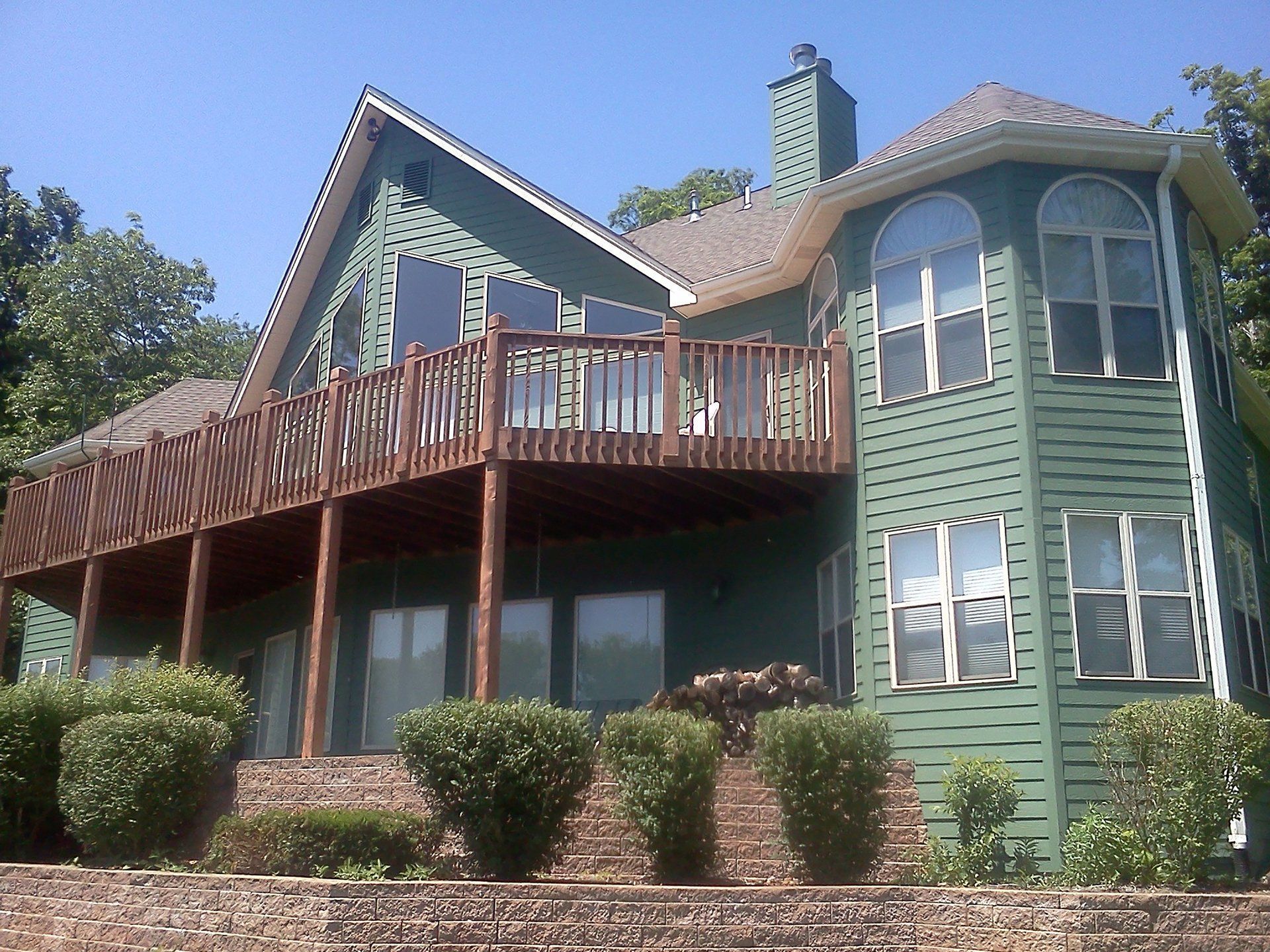 Green two-story house with a wooden deck, arched windows, and shrubs in front