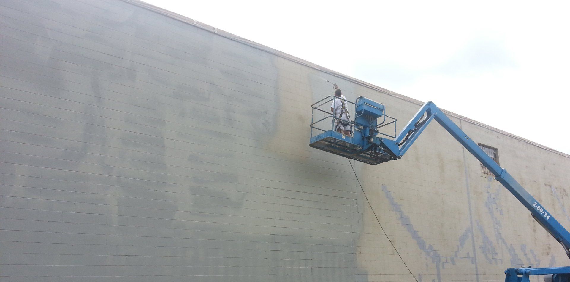 Worker in a blue lift cleaning a large concrete wall with visible water stains.