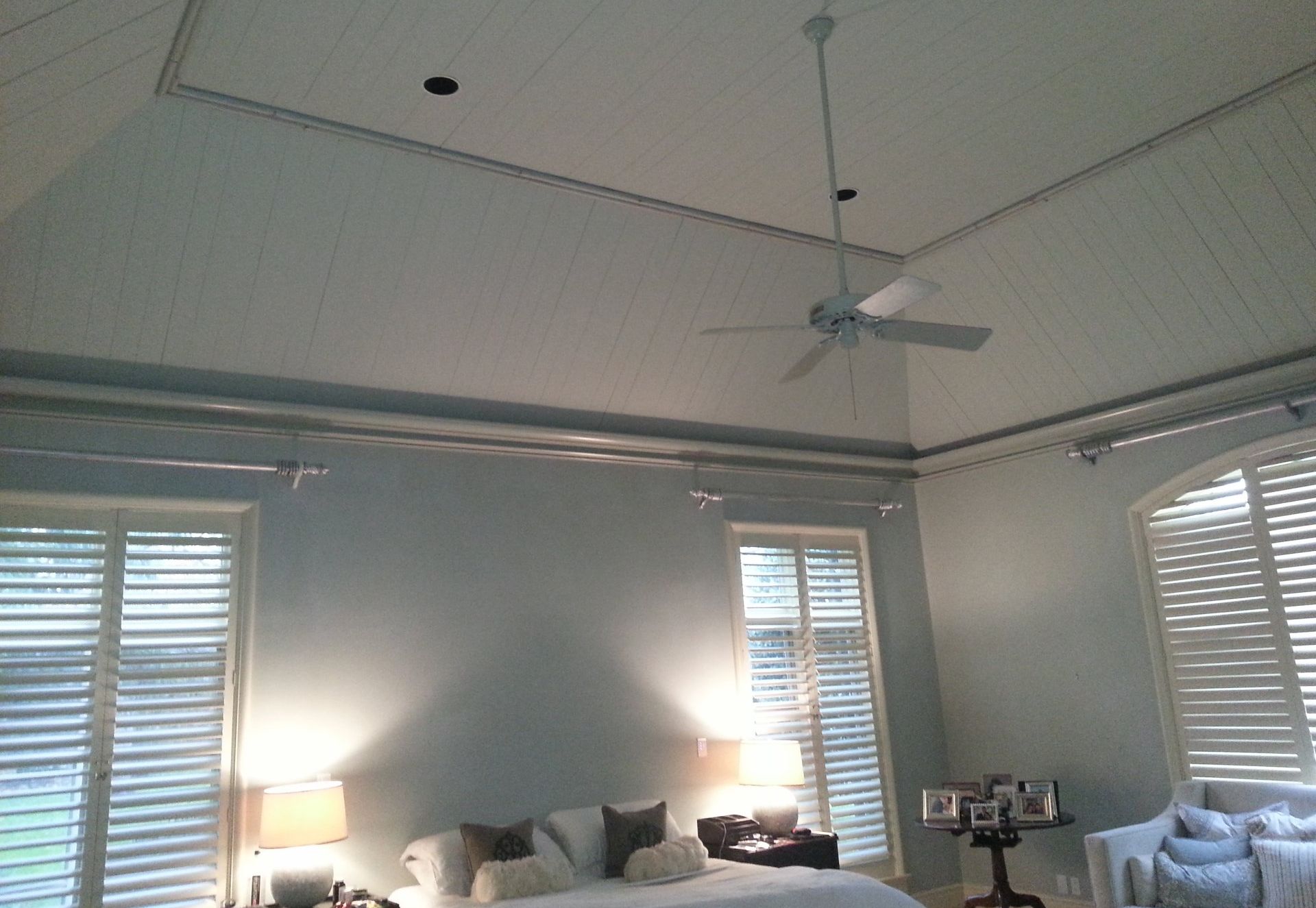 Bright bedroom with high ceiling, ceiling fan, windows with blinds, and bedside lamps illuminating the room