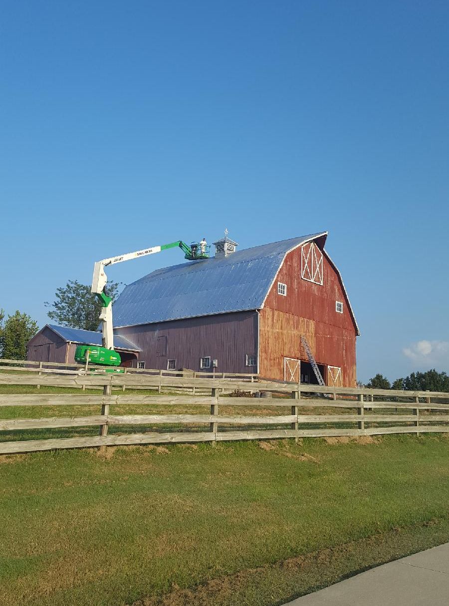 Red barn with a green cherry picker beside it, behind a wooden fence under a clear blue sky.