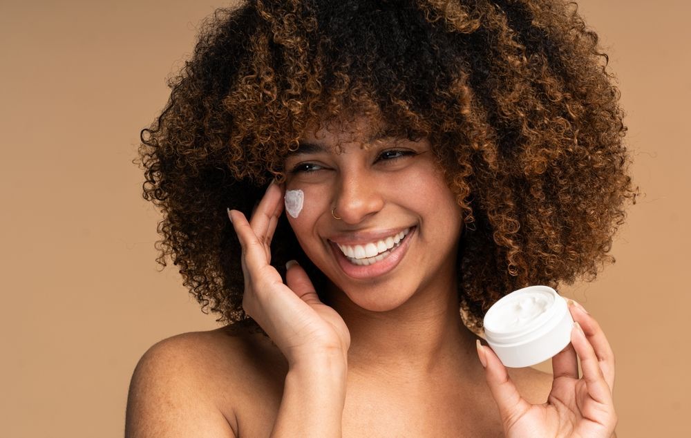 Smiling person applying cream to face, holding a small jar against a beige background.