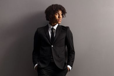 Person in a black suit and tie standing against a gray background, hands in pockets, looking to the side
