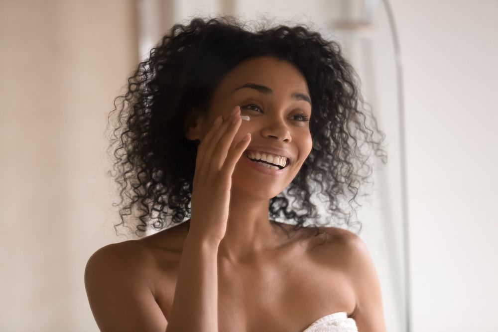 Smiling person with curly hair applying skincare in a softly lit bathroom