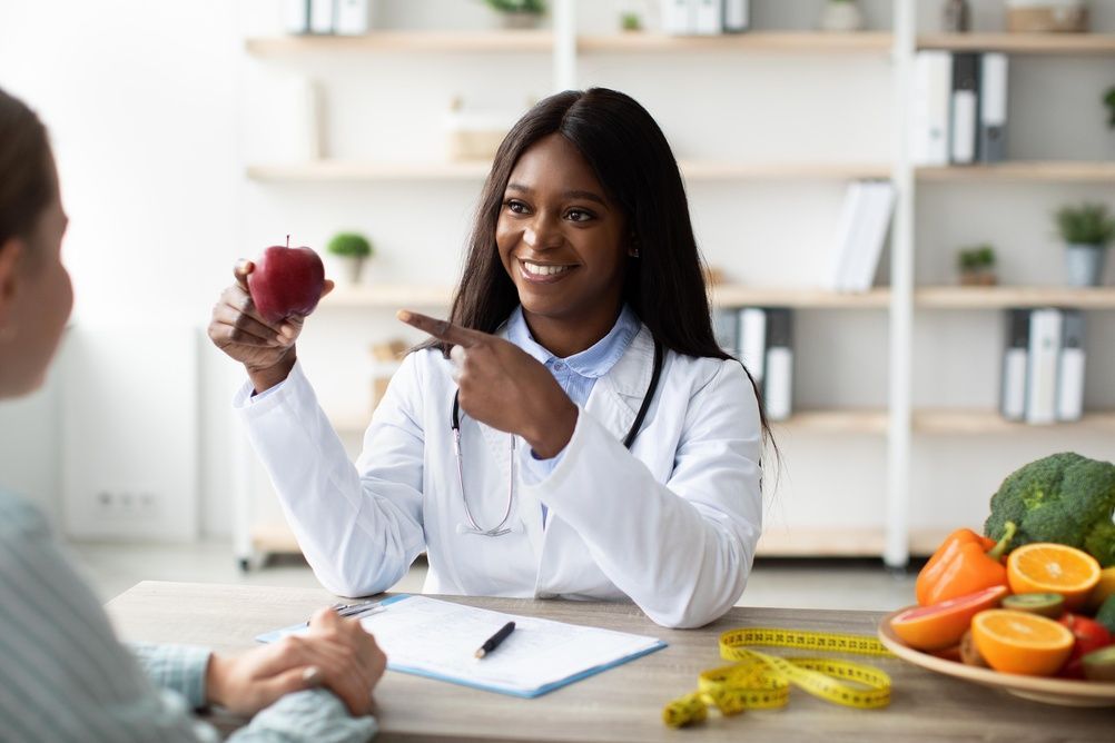 Doctor holding a red apple and pointing to a patient during a nutrition consultation with fruits on the desk