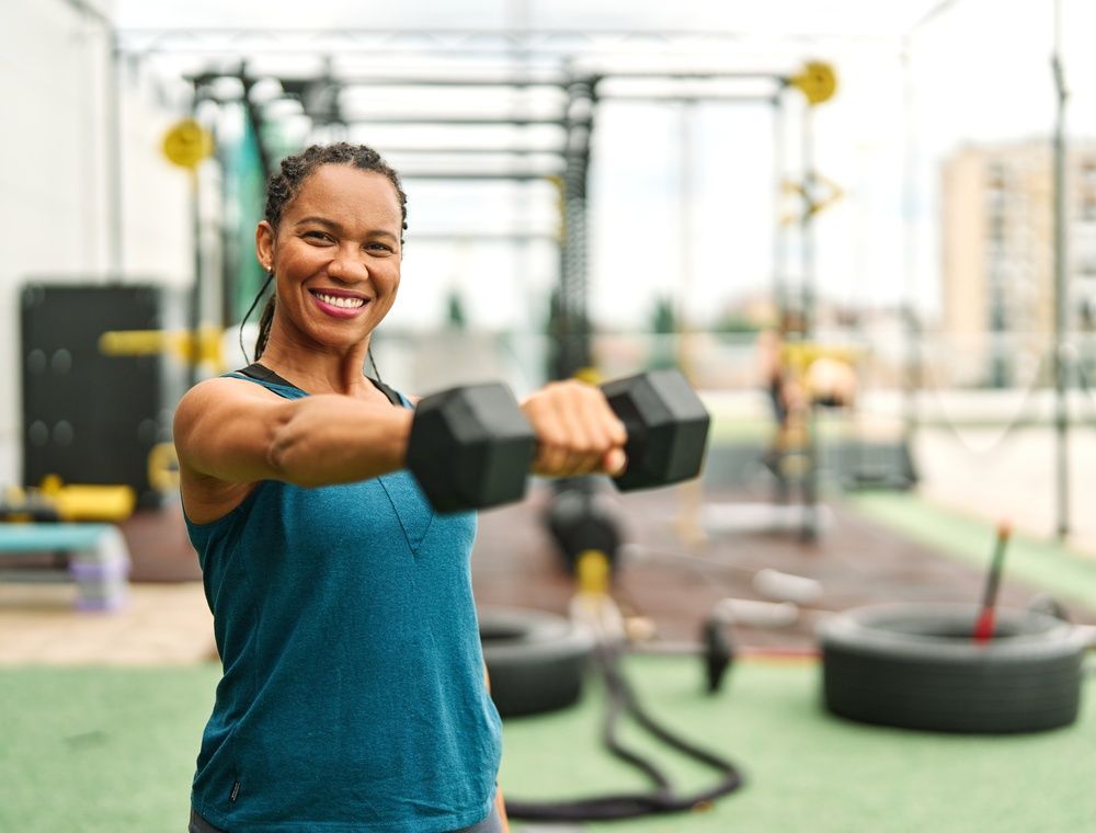 Woman smiling and holding dumbbells out in a gym, with exercise equipment in the background
