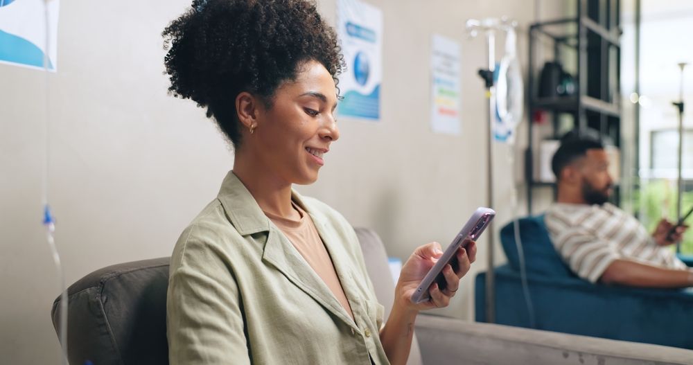 Woman smiling at her phone in an office, with a coworker blurred in the background.