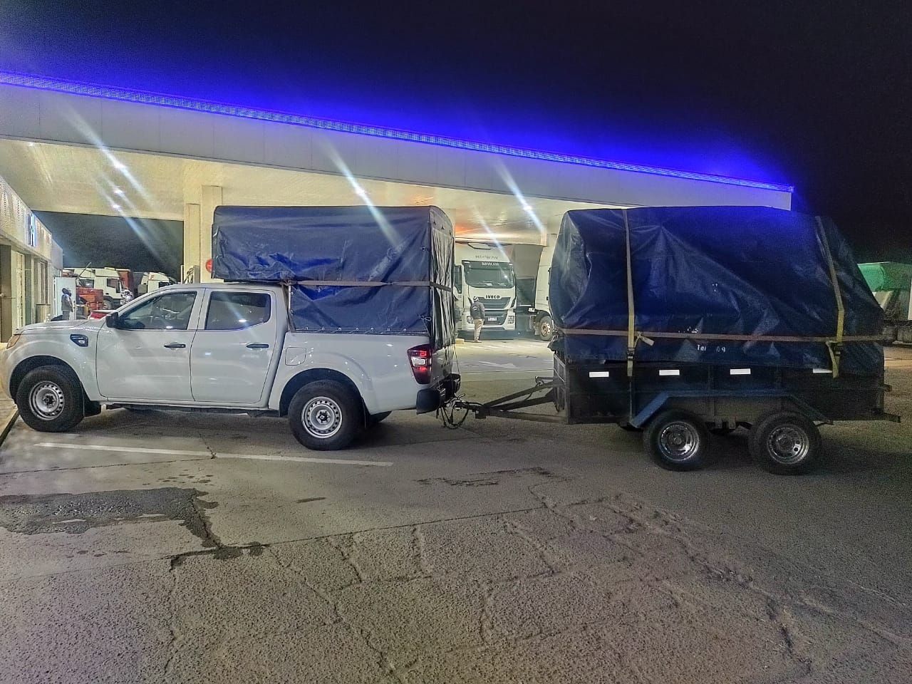 Una camioneta blanca que arrastra un remolque, ambos cubiertos con lonas azules, estacionada en una gasolinera por la noche.