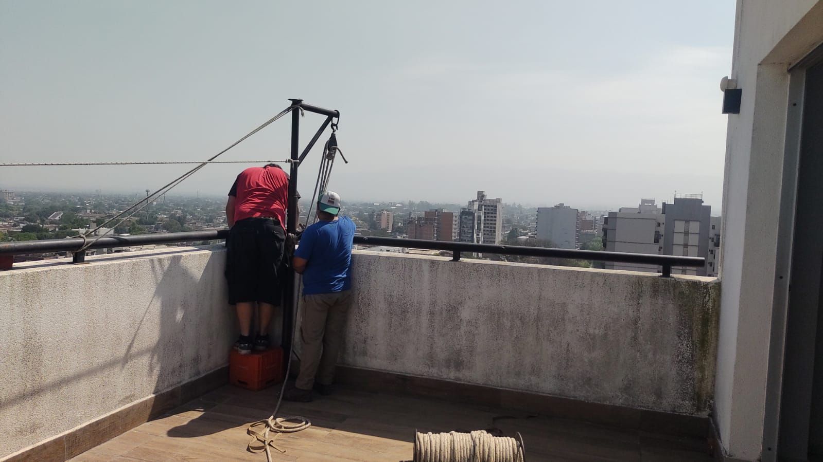 Dos personas en un tejado, operando un sistema de cables para bajar algo. El horizonte de la ciudad al fondo.