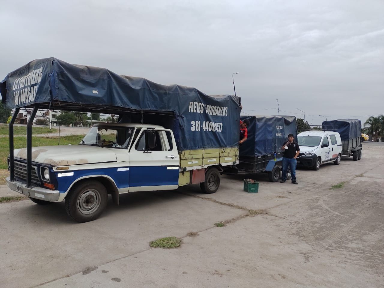 Camiones con lonas azules estacionados, con gente cerca. Cielo gris.