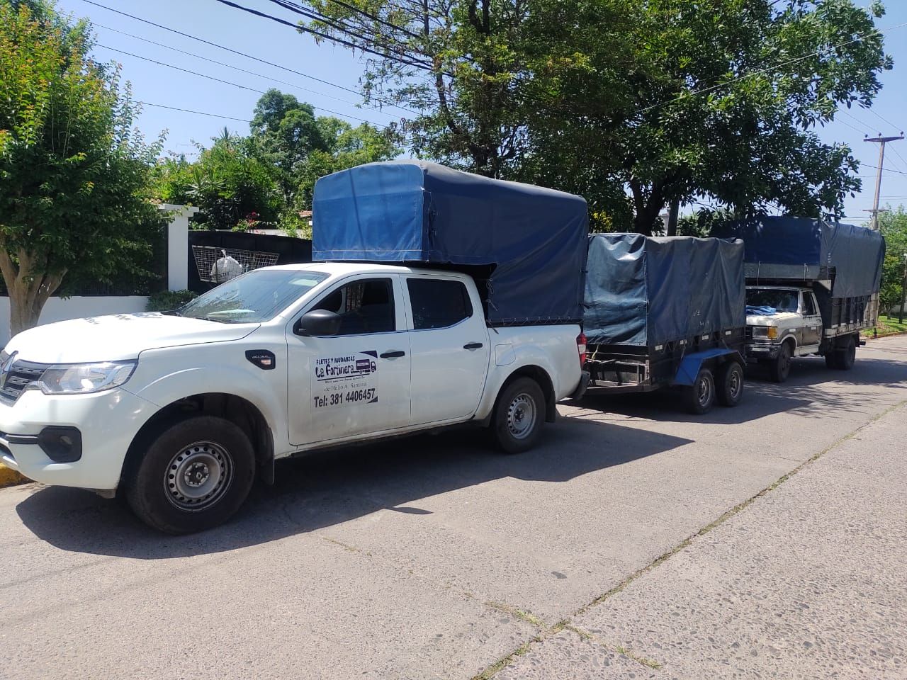 Camioneta blanca que arrastra dos remolques, todos con cubiertas de lona azules, estacionados en una calle.