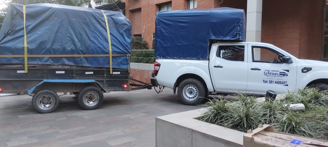 Una camioneta blanca arrastrando un remolque, ambos cubiertos con lonas azules, estacionada afuera de un edificio.
