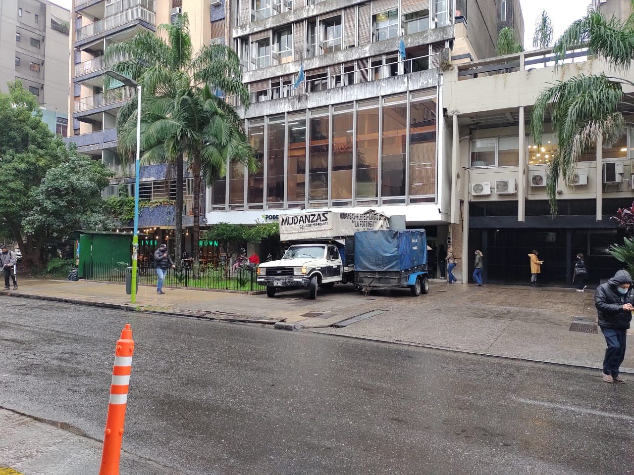 Escena callejera con un edificio, camiones estacionados y gente. Cielo gris y carretera mojada.