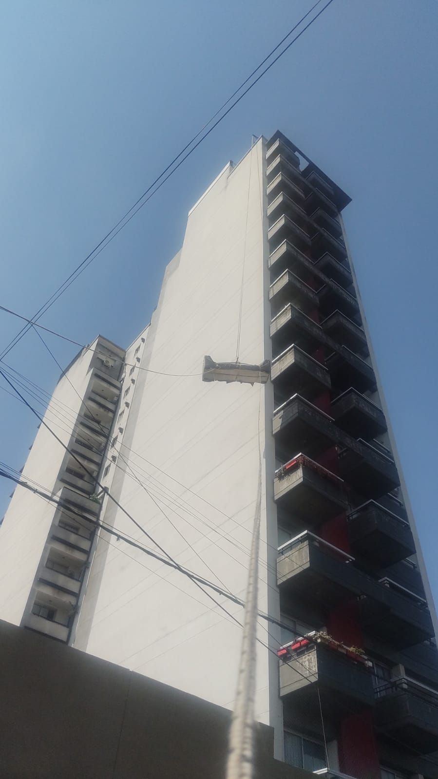 Edificio de apartamentos alto y blanco con balcones, cables y un cielo azul claro.