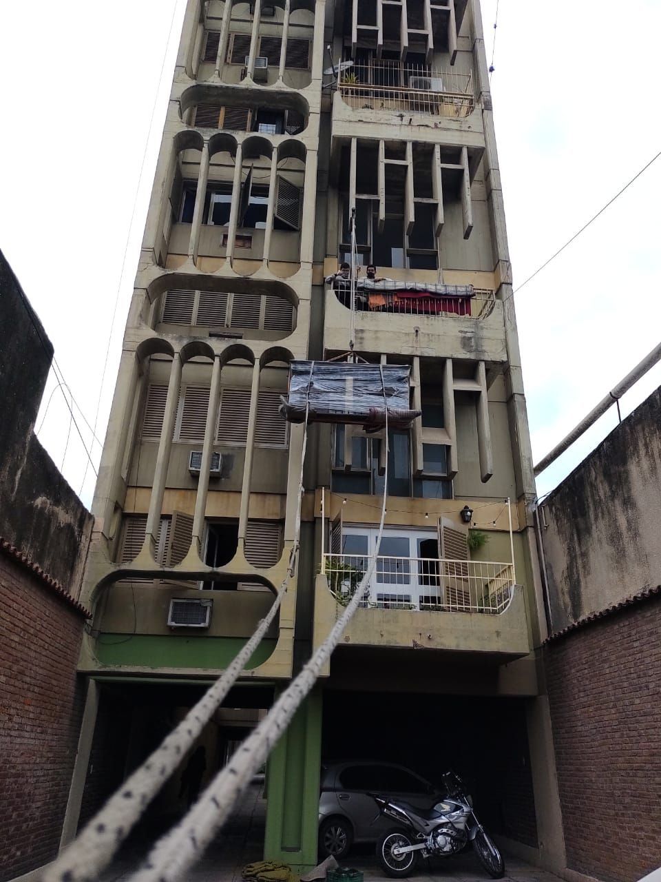 Edificio alto y desgastado de hormigón con ventanas y balcones empotrados; coche y motocicleta aparcados debajo.