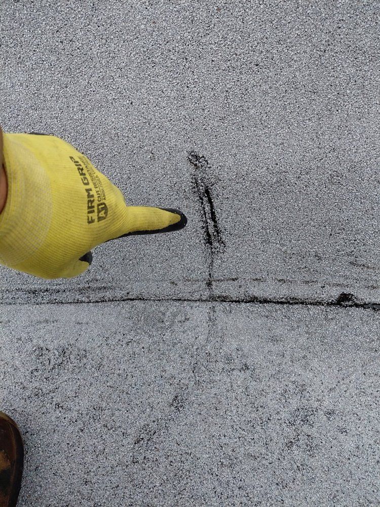 Gloved hand points to a crack in a gray, textured roof surface.