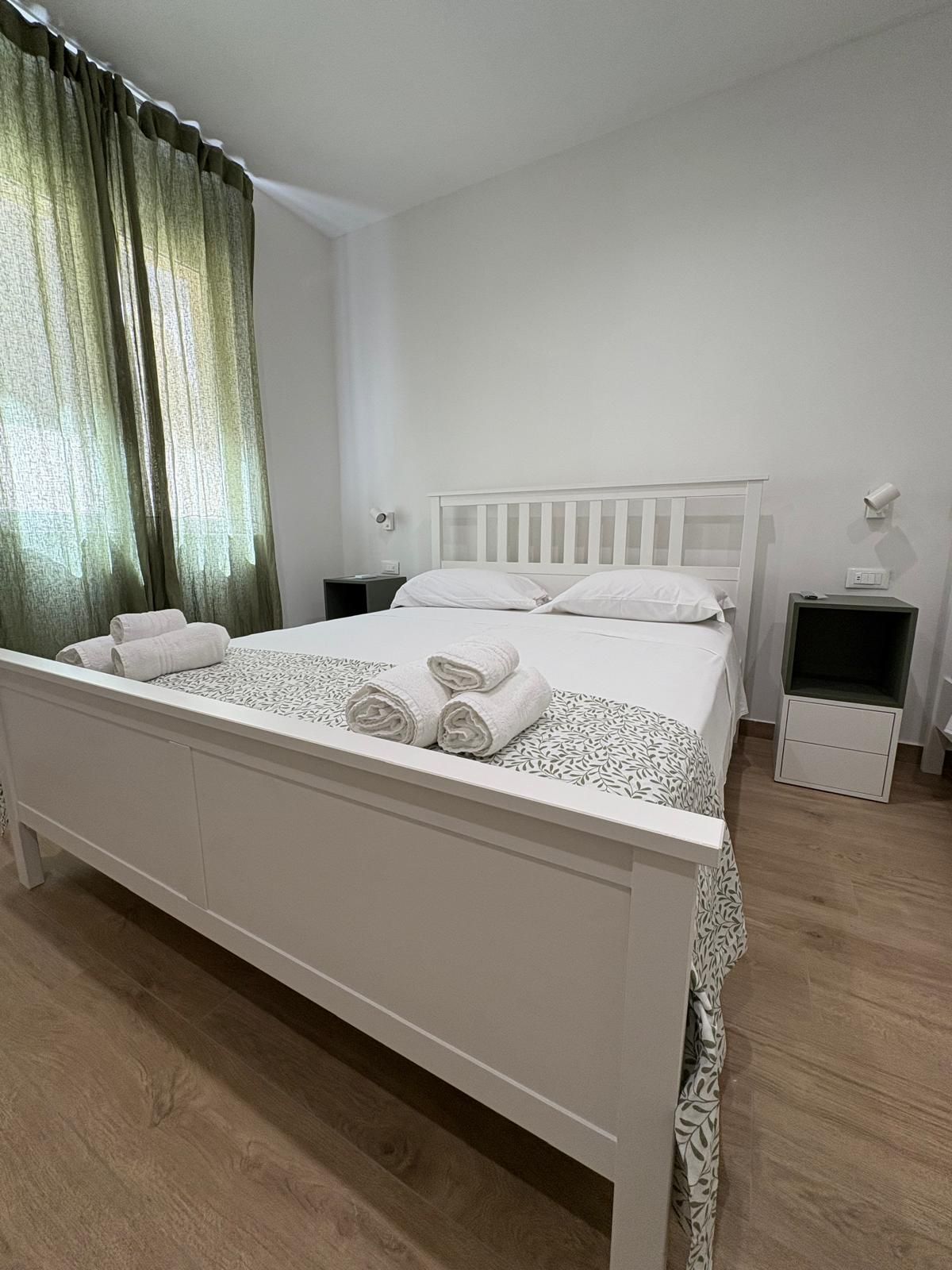 Camera da letto bianca con letto, comodini e tende verdi.