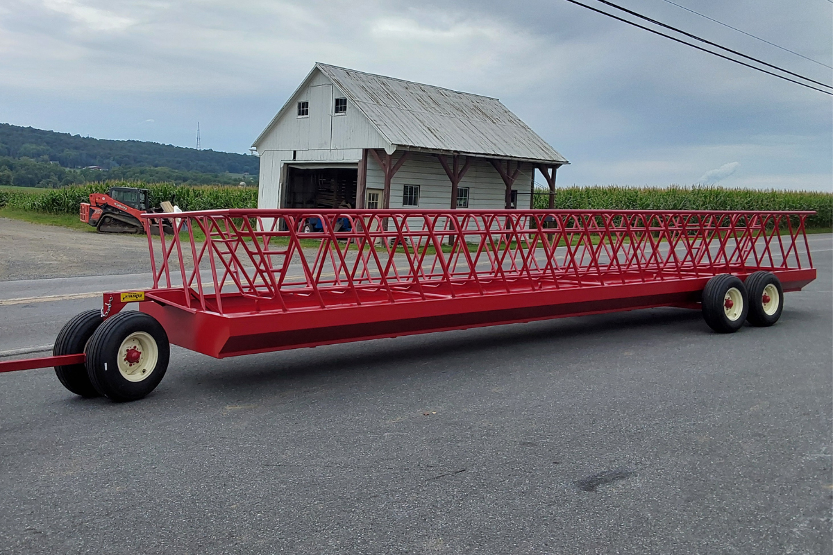 Heavy-duty red double-wide feeder wagon with an open metal hay-feeding basket, sturdy steel frame, and tandem wheels, designed for feeding livestock and cattle on the farm.
