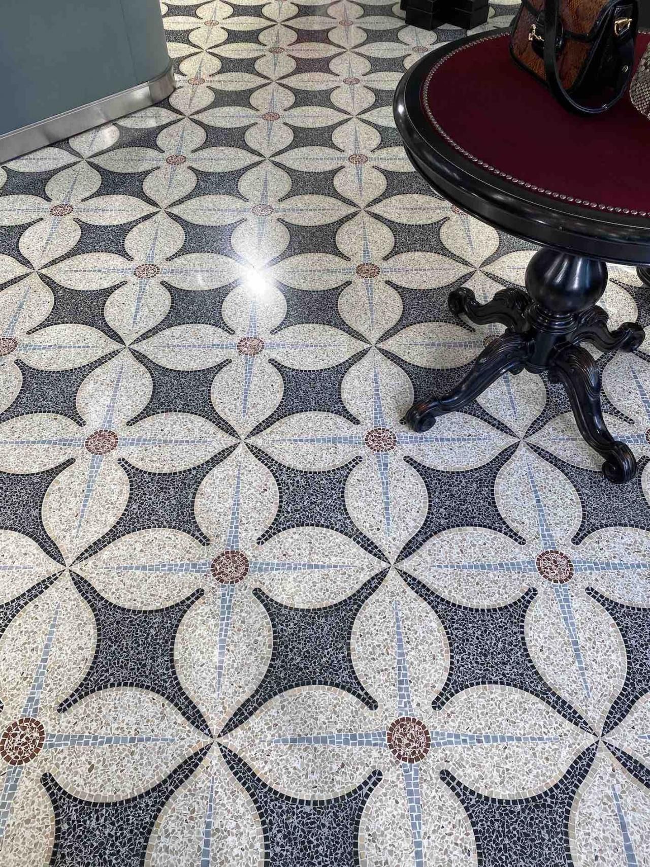 Vista dall'alto di un pavimento decorativo a mosaico con un motivo ripetuto a quadrifoglio grigio chiaro e scuro, accanto a un tavolino.
