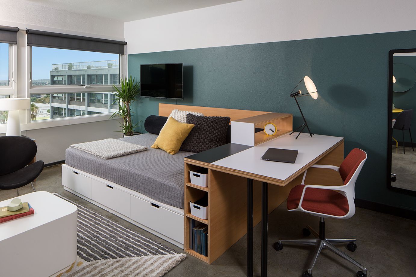 Modern studio apartment with bed, desk, wall-mounted TV, and large window with city view