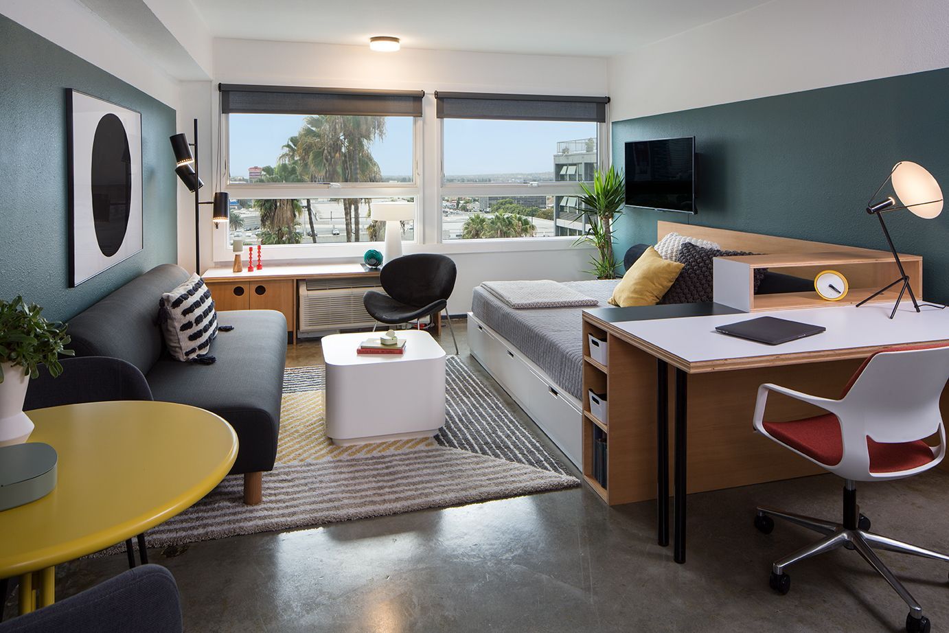 Modern studio apartment with bed, sofa, desk, window view, and wall-mounted TV