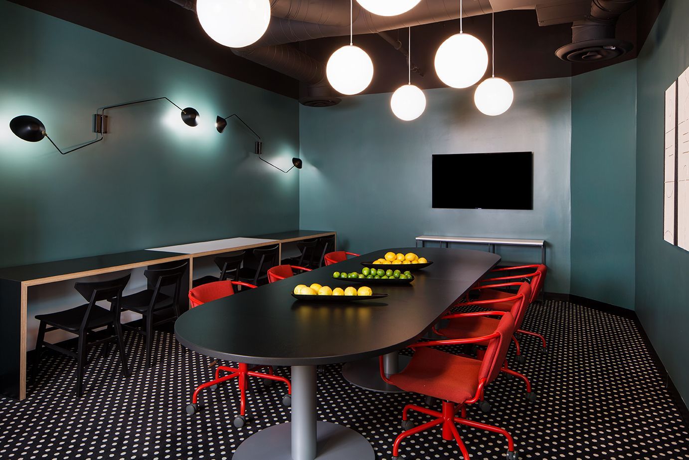 Modern conference room with red chairs, oval table, wall-mounted screen, and hanging globe lights