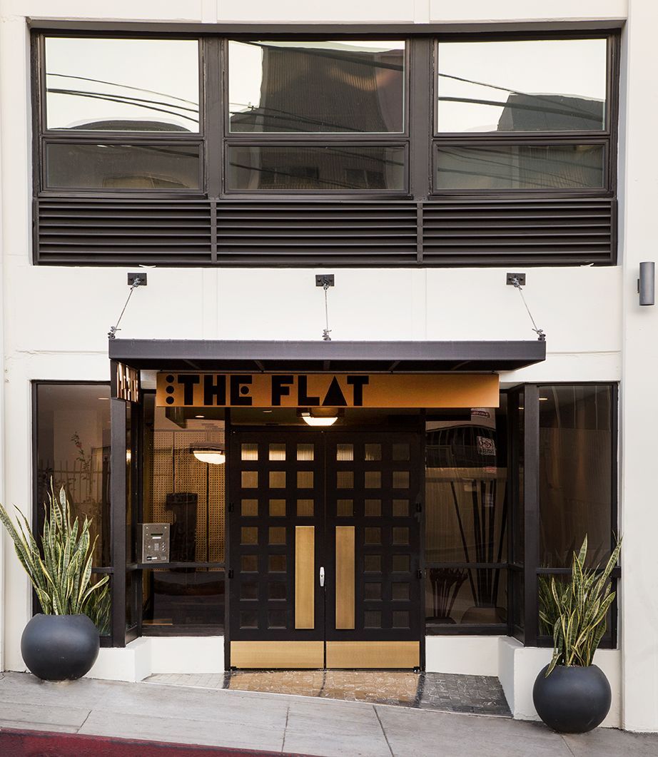 Storefront entrance with sign “THE FLAT,” black awning, and potted plants at the doorway