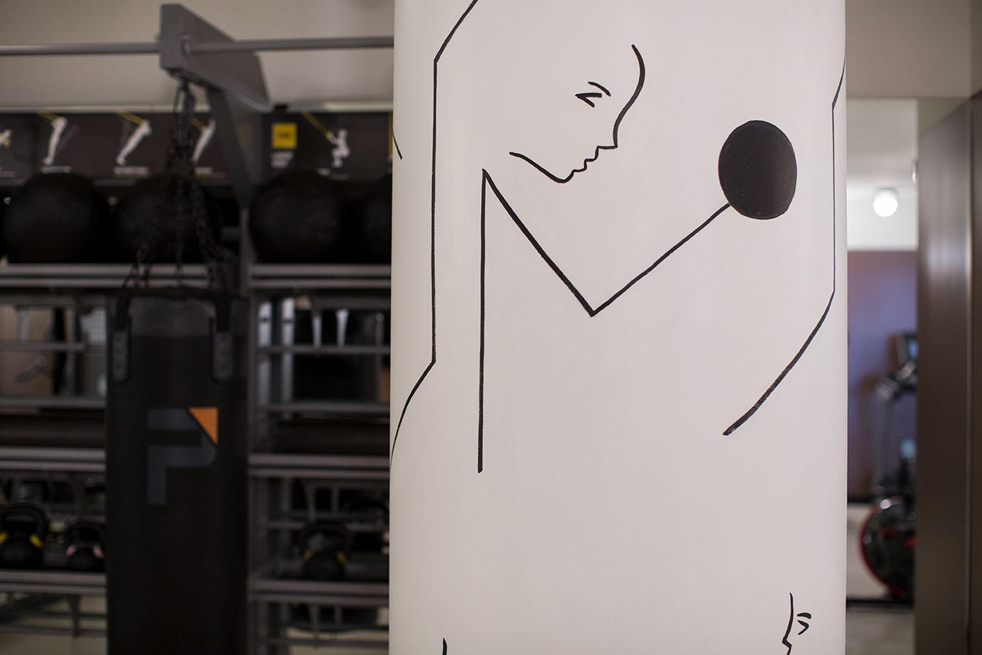 Graffiti-style black line drawing on a white pillar in a gym, with equipment blurred in the background