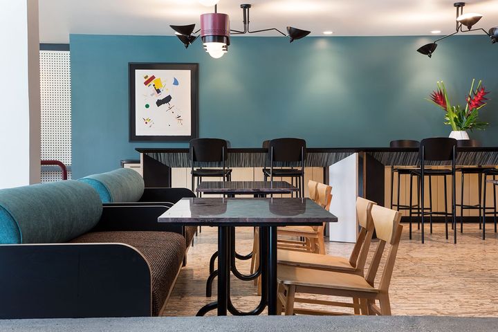 Modern lounge with blue couch, black tables, high stools, teal walls, and framed art; no people