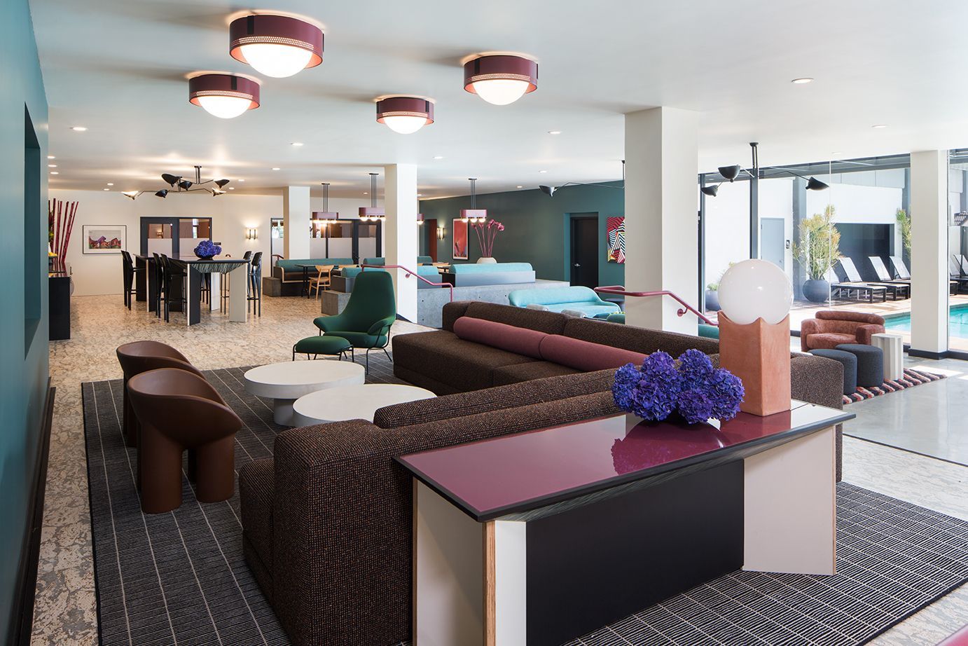 Modern hotel lounge with brown sofas, patterned rugs, and red pendant lights in a bright open space