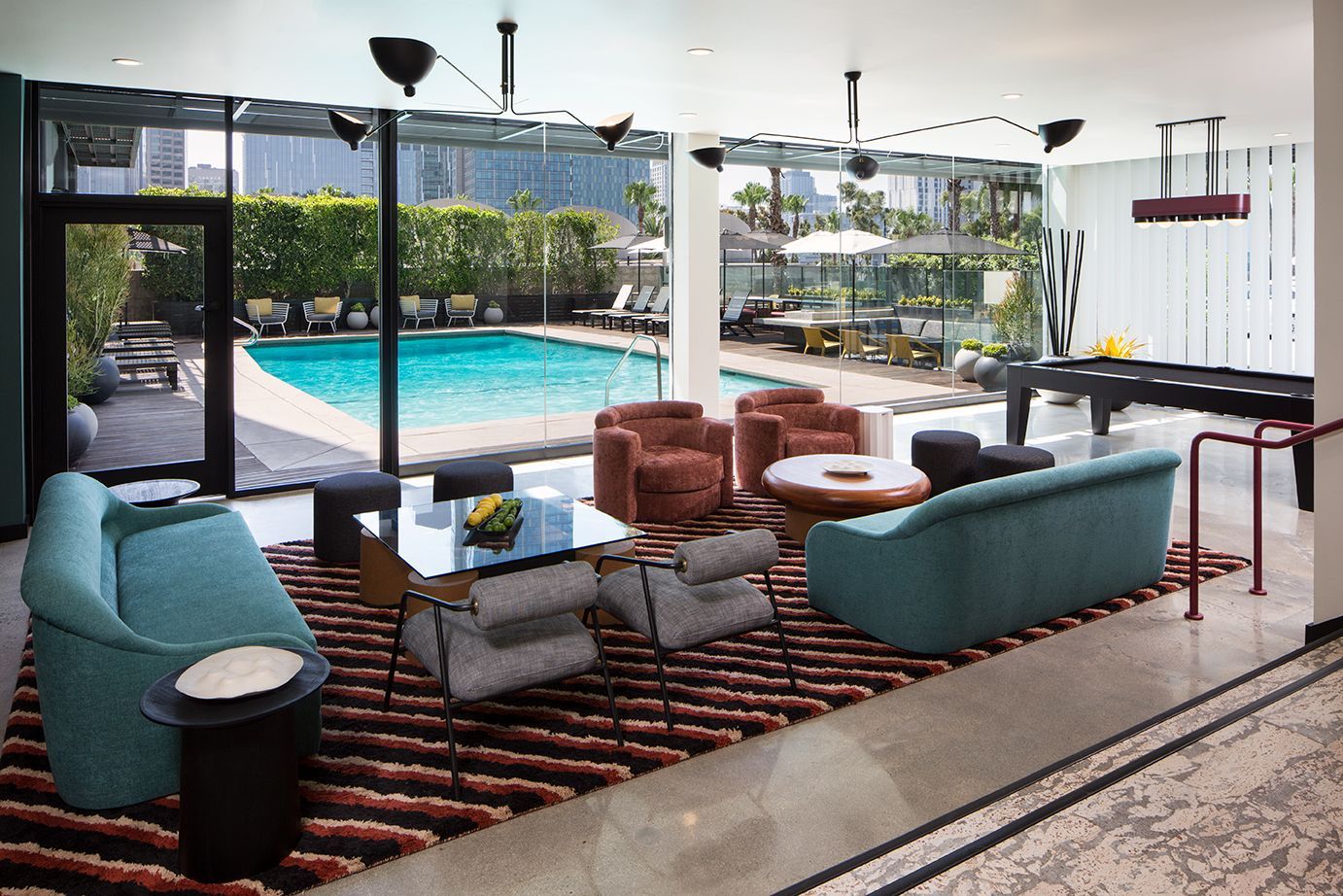 Modern indoor-outdoor lounge with turquoise sofas, striped rug, and pool view through glass walls
