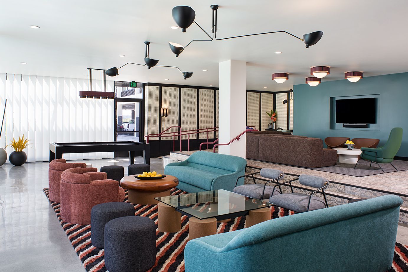 Modern lounge with teal and brown sofas, glass tables, and a wall-mounted TV in a bright open room