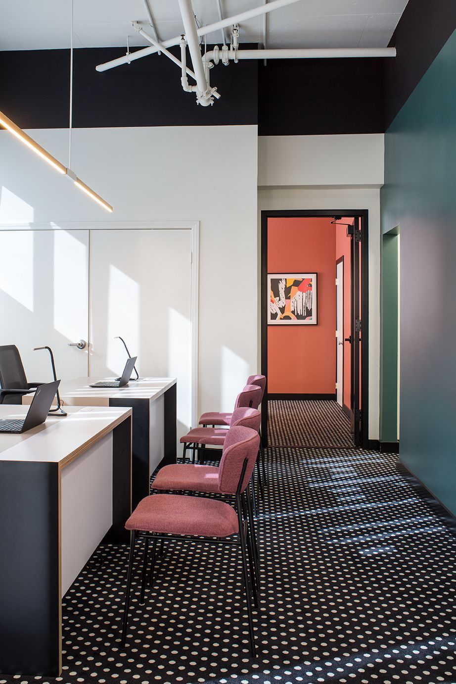 Modern office hallway with desks, pink chairs, patterned carpet, and an open doorway to a coral room