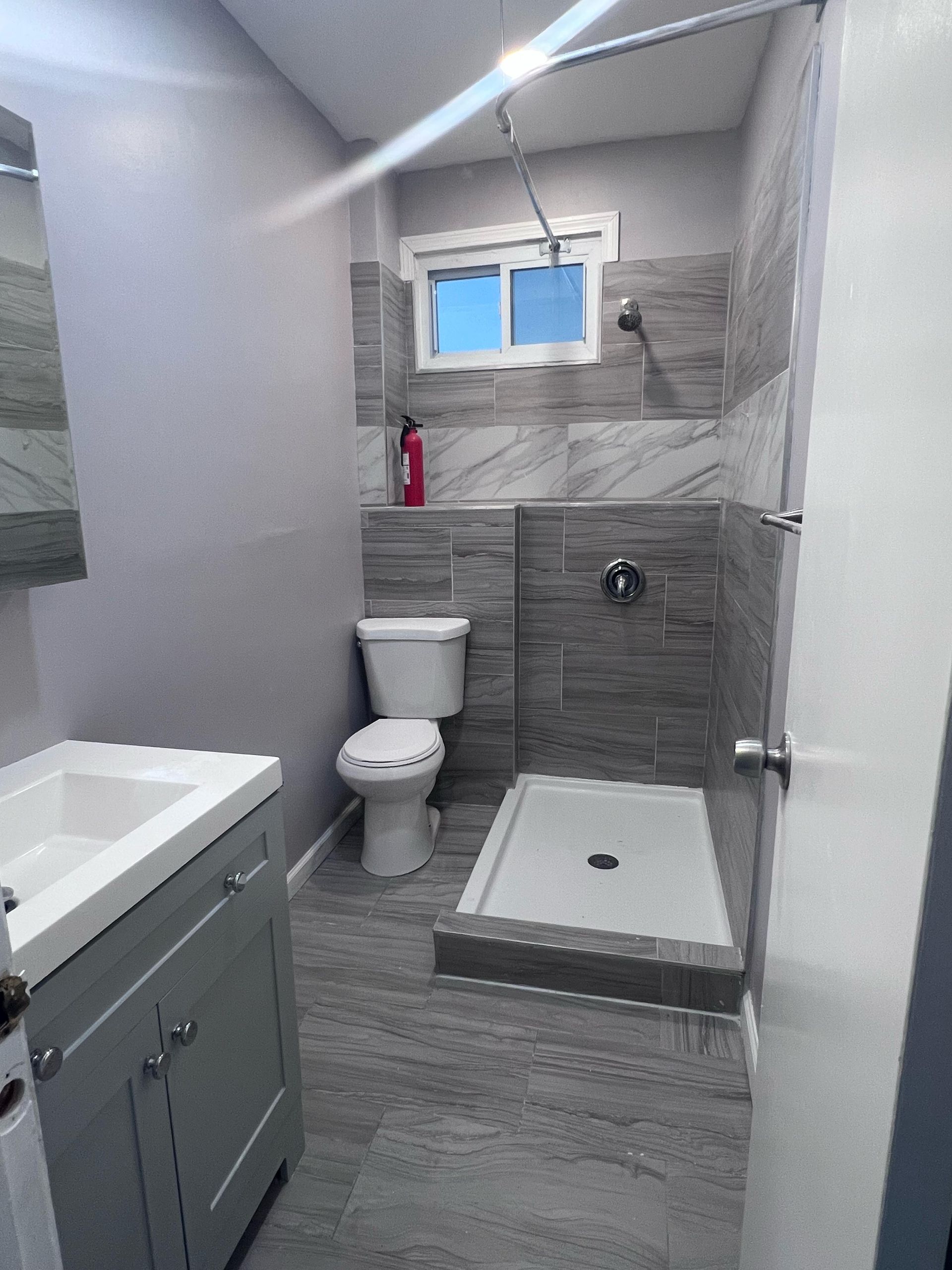 Small gray bathroom with toilet, sink, shower, and window above the shower.