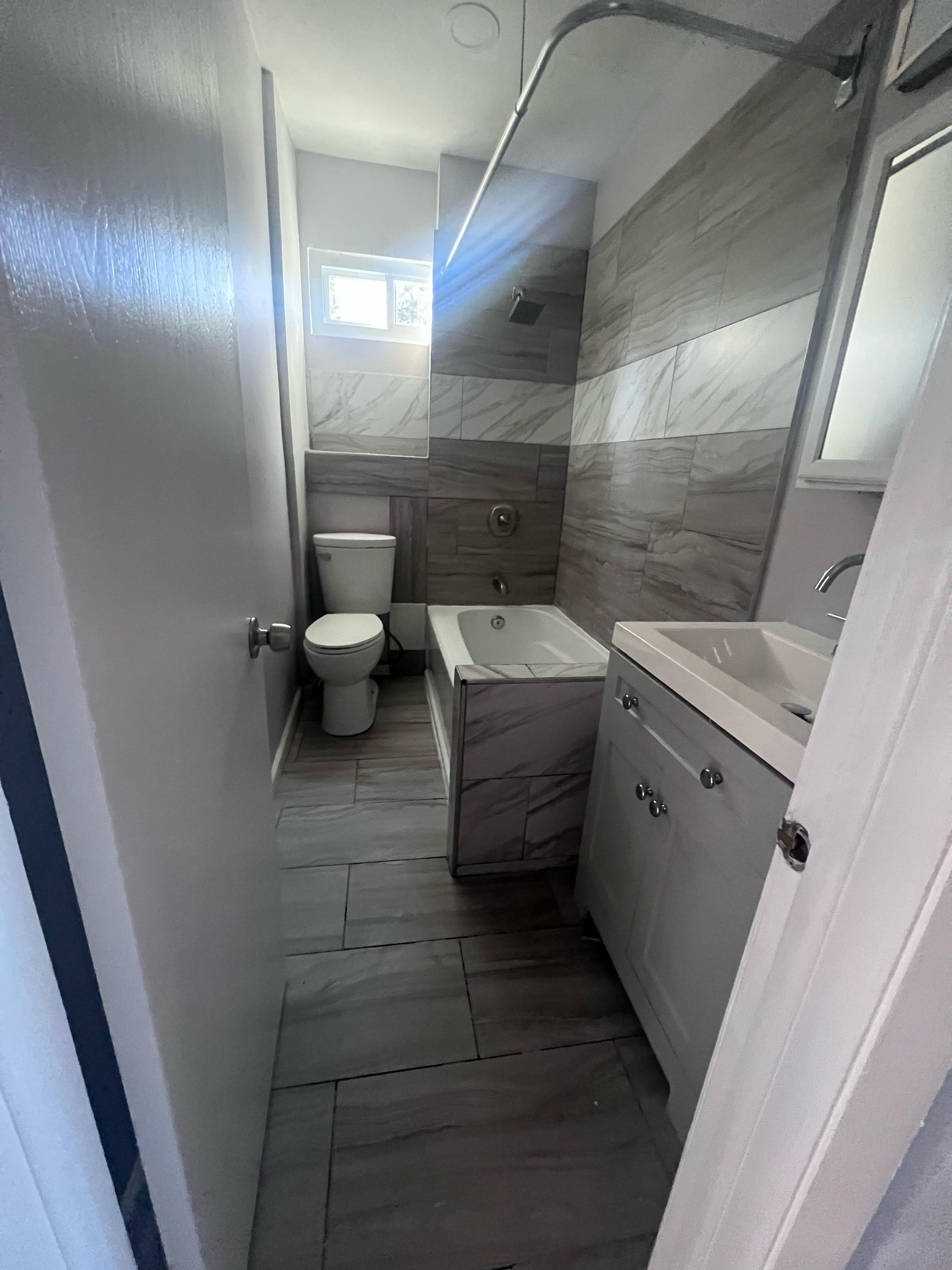 Narrow bathroom with toilet, shower, sink cabinet, and gray tile walls and floor.