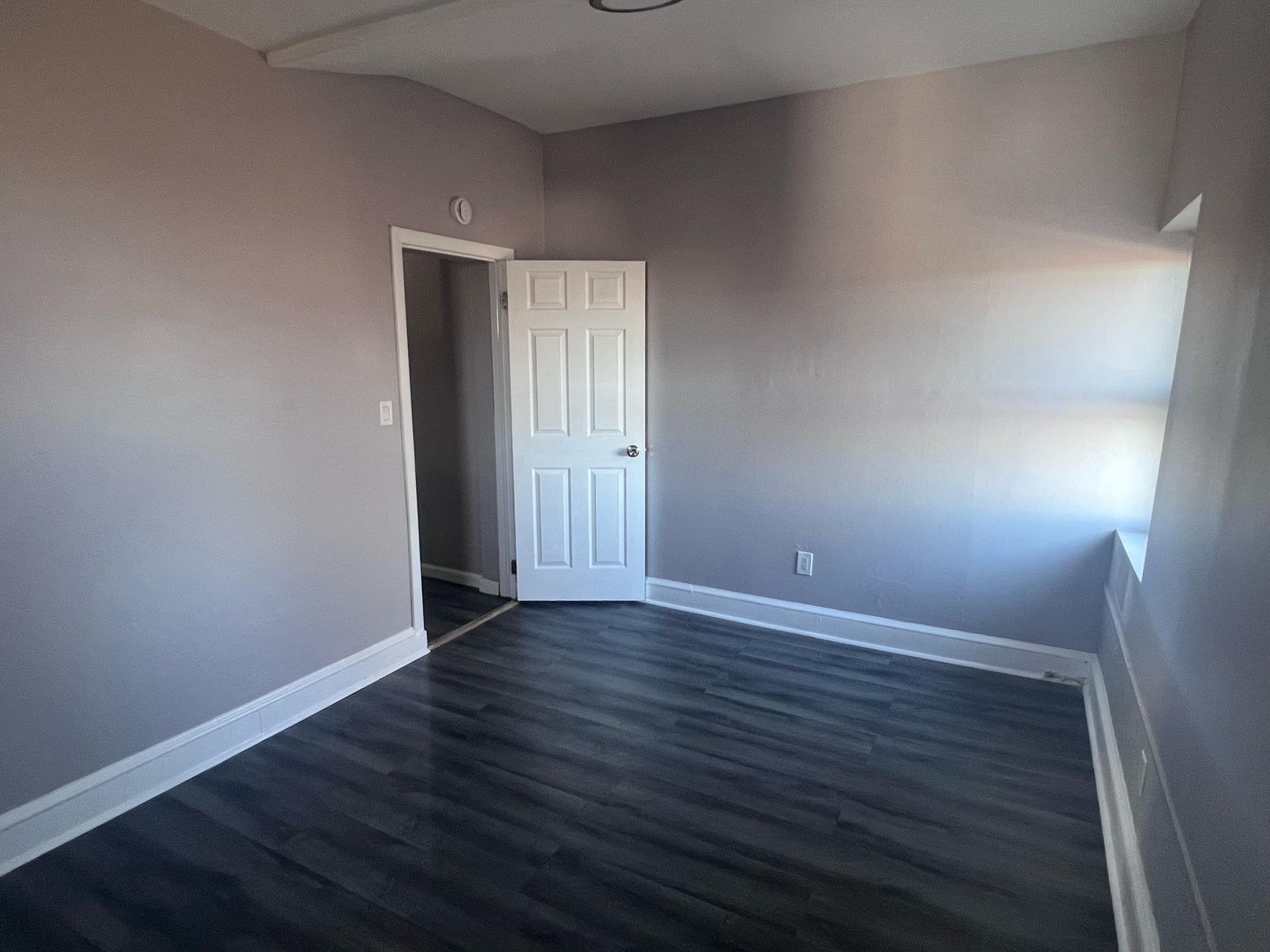 Empty gray bedroom with dark wood flooring, open door, and sunlight on the right wall